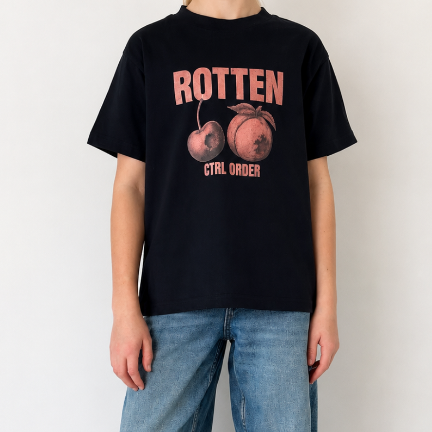 A person wearing a navy graphic T-shirt with pink and orange text and fruit imagery, paired with loose blue jeans and black-and-white sneakers, standing against a plain white wall.