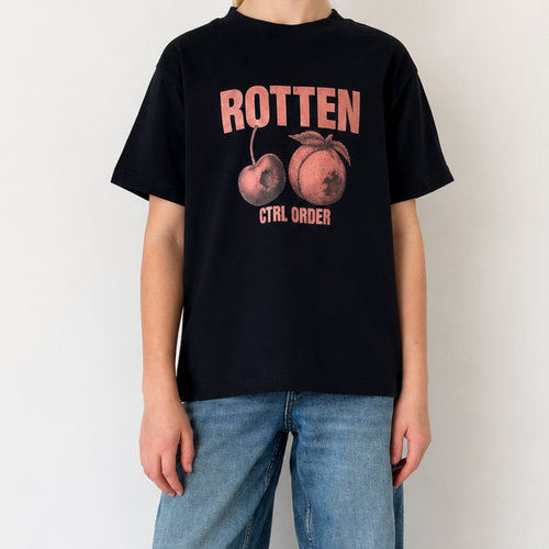 A person wearing a navy graphic T-shirt with pink and orange text and fruit imagery, paired with loose blue jeans and black-and-white sneakers, standing against a plain white wall.