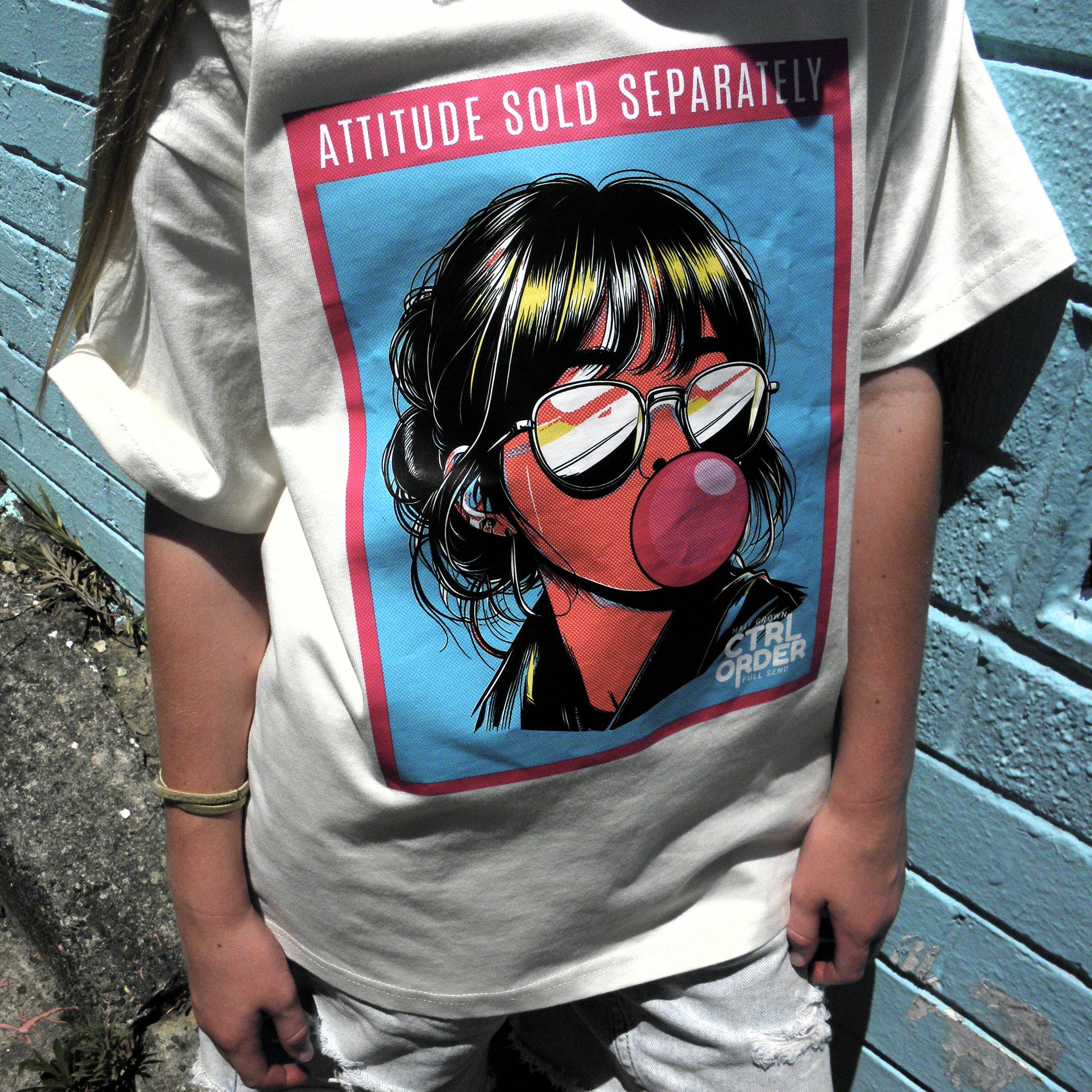 Close-up of an ecru t-shirt featuring the ‘Attitude Sold Separately’ graphic, worn by a kid against a blue brick wall.