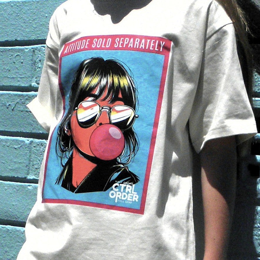 Kid wearing an ecru t-shirt with ‘Attitude Sold Separately’ graphic featuring a girl blowing a pink bubble gum bubble.